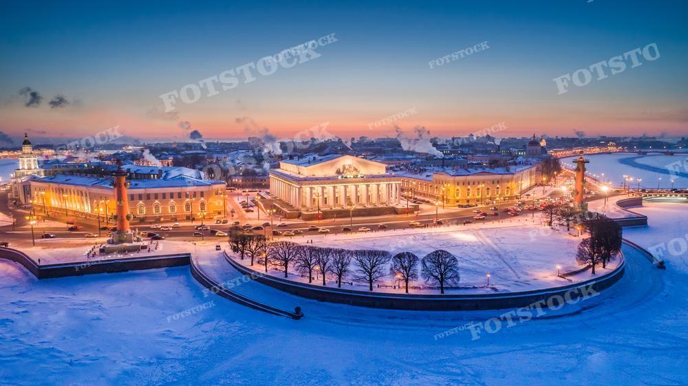 На фотографии изображён зимний Санкт-Петербург с высоты птичьего полета. Видны заснеженные набережные и здания, подсвеченные уличными огнями. В центре кадра расположен Стрелка Васильевского острова: здание Биржи и Ростральные колонны. Это фото может использоваться для иллюстрации путеводителей, культурных и исторических статей, образовательных материалов, а также в качестве изображения для туристических сайтов и блогов, посвящённых России и Санкт-Петербургу.